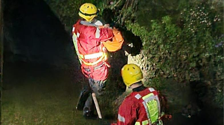 Two firefighters wading through a shallow river in Newton Abbot. The firefighters are wearing red and yellow rescue flood suits. 