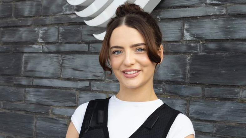 Olivia Browne yn sefyll o flaen wal y Senedd, yn gwisgo fest rhedeg