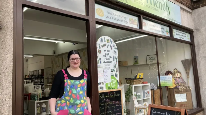 A woman with black hair and glasses wears a black top and a colourful apron with leaves on it as she stands in the doorway of her refill shop. Large bottles of liquid with big pumps can be seen inside the shop and there are scarecrows, shelving, and signs in the window.