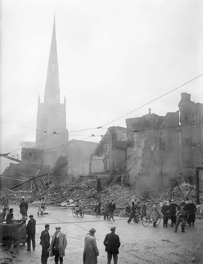 A black and white image of people in the streets next to partially and fully destroyed buildings and piles of rubble from which smoke rises. A church spire can be seen in the background.
