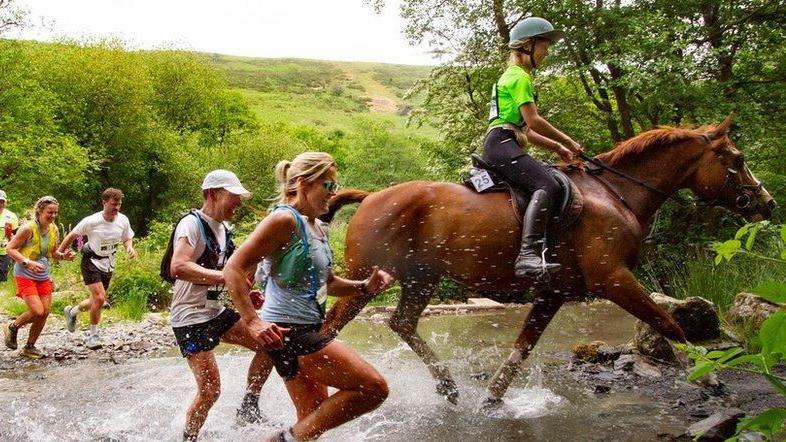 Two women running next to a horse through a small stream surrounded by fields and treea