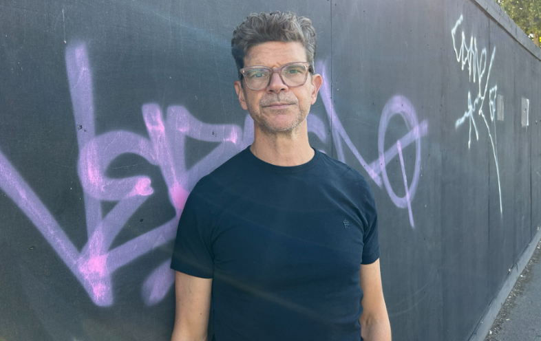A man with grey-black hair looks at the camera. He is bespectacled and is wearing a blue T-shirt