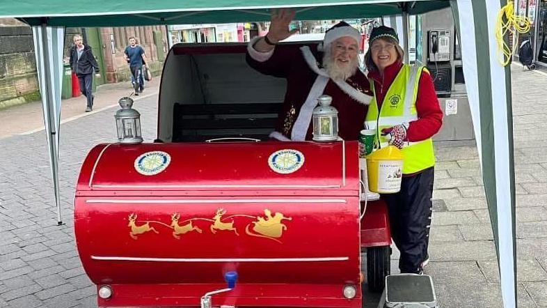 A red sleigh with Santa in on the main high street in Belper. Next to the sleigh is a woman wearing a hi vis jacket, with a bucket collecting money.  