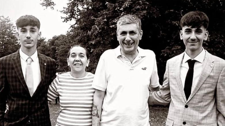 A black and white image shows two young men with dark hair wearing suits and stood on either side of their parents, with the mother wearing a striped t-shirt and the father a polo shirt, with trees in the background.