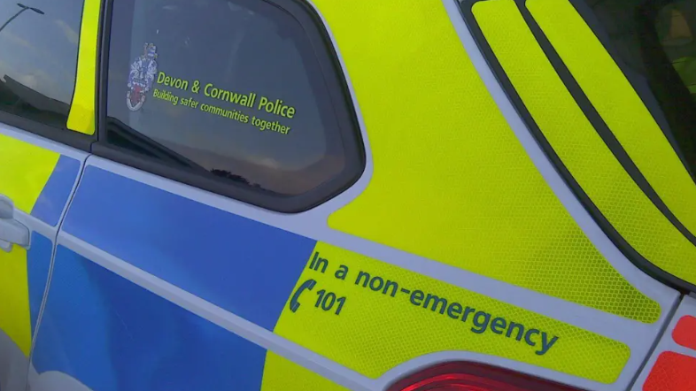 The side of a police car which has blue and yellow stickers on the side of the car. The back window says 'Devon & Cornwall Police Building safer communities together'. One of the back yellow panels also advises people to call 101 in a non-emergency.