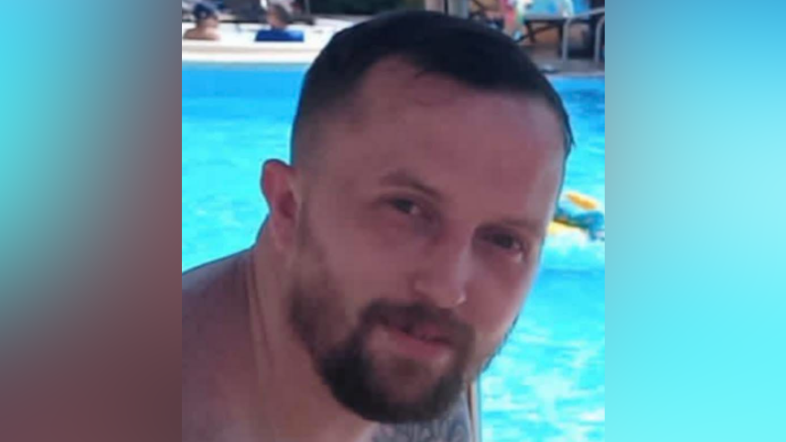 A photo of Stephen Holmes. He is shown from the shoulders up sitting in front of a pool. He has short brown hair, and a beard. He is smiling at the camera.