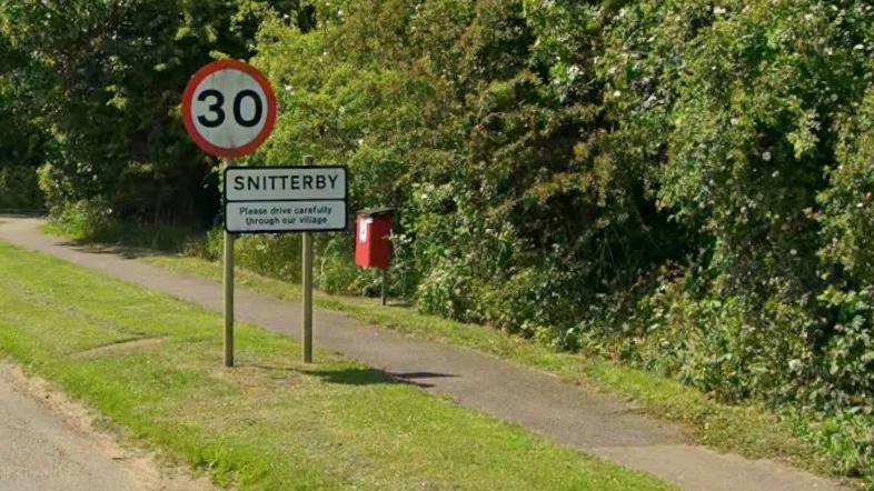 A village sign - rectangular and white - held up by two posts on a grass verge next to a footpath and green shrubbery. The sign has black text reading "Snitterby: Please drive carefully through our village". A 30-mile per hour speed limit sign stands above the village sign. It is circular, with a red border and the black number 30 on a white background. A red bin for dog waste stands nearby.