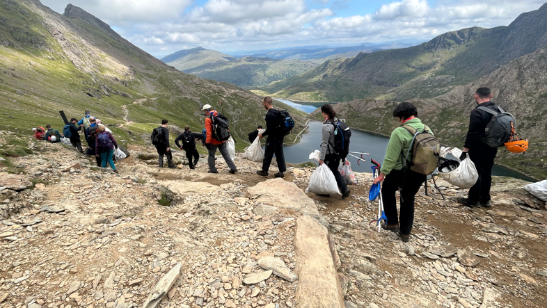 A large group of people trekking on a path with mountains behind them. They are all carrying bags of rubbish and litter pickers.