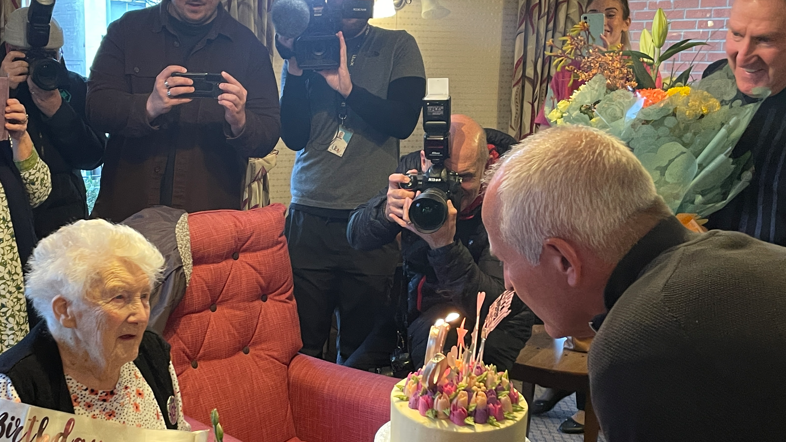 Joan is sat in a chair with Steve Bull and Steve Daley giving her a pretty iced cake topped in pink and purple flowers and candles. There are people looking on with cameras to capture the moment.