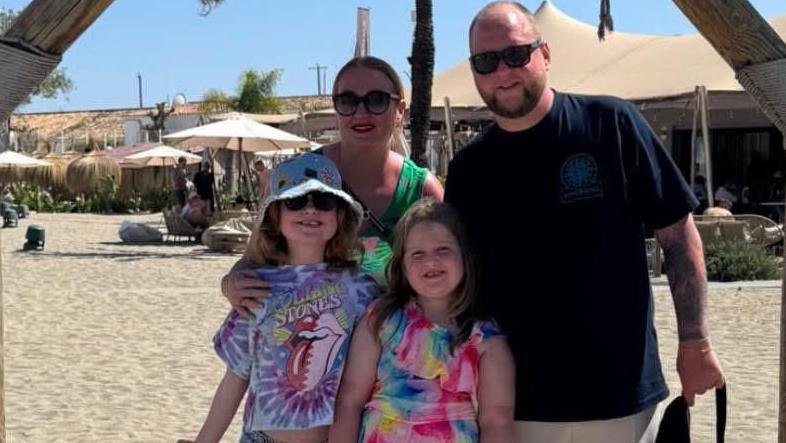 A family of four - a man, a woman and two young girls - stand on a beach in a hot destination. They are wearing T-shirts and sunglasses, and one of the girls is wearing a bucket hat. They are all smiling at the camera.