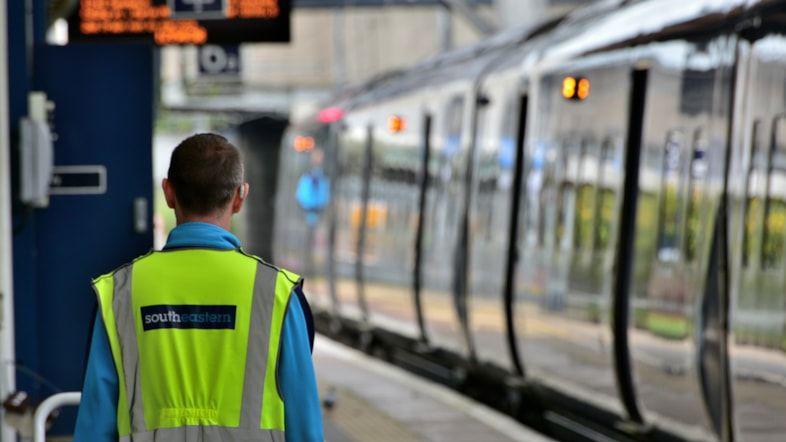 A member of station staff wearing hi-vis watches a Southeastern train pull into a station.