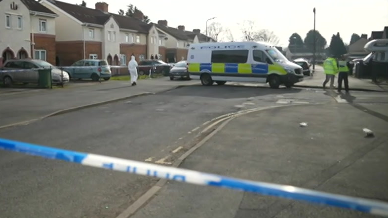 Police tape can be seen in the foreground with a police van and officers standing nearby in the road