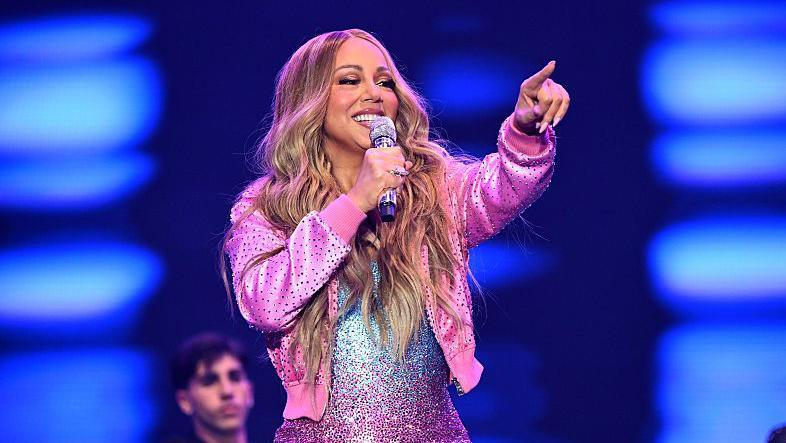 Mariah Carey in a blue, silver and purple dress which is sparkly. She is wearing a pink jacket and is smiling and pointing at the crowd while holding a micrphone.