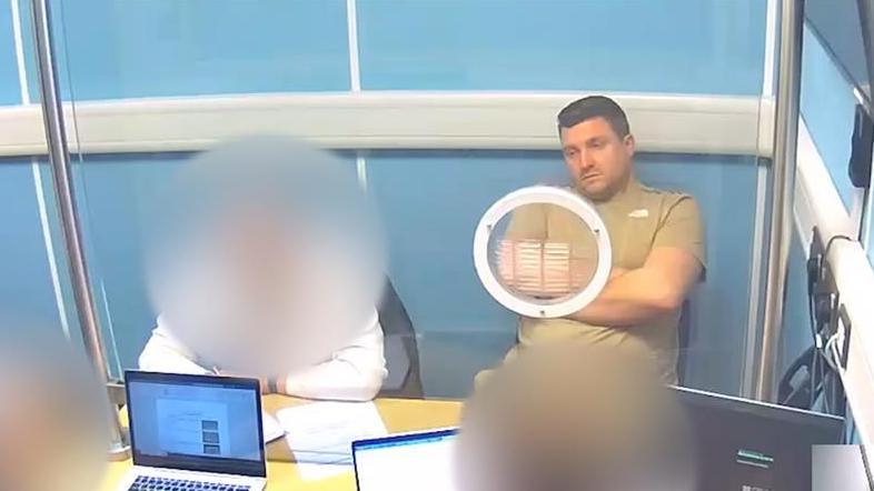 A man in police custody wearing a brown top is being questioned by two detectives who are sat in front of him, both using laptop computers. Alongside the arrested man is a man wearing a white shirt and his face is blurred out 