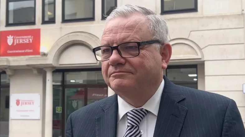 Constable Richard Vibert is wearing a black pin stripe suit with a white shirt. He is wearing glasses and is stood outside the Government of Jersey building on Broad Street