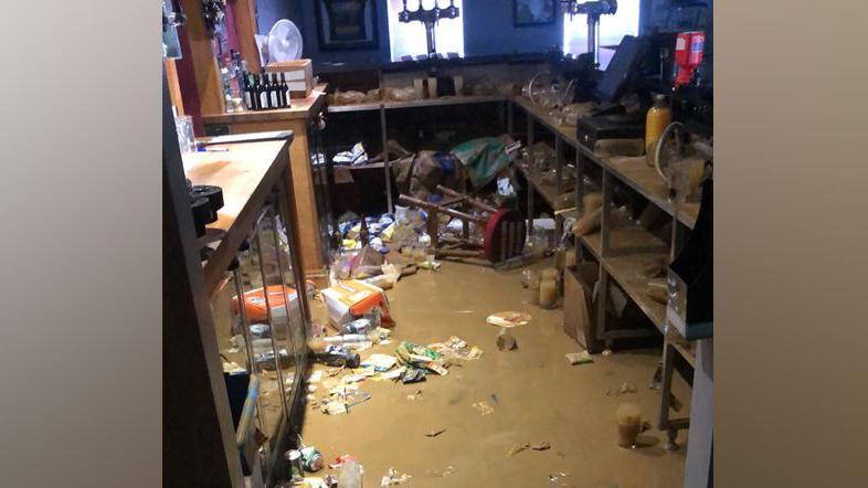 Flood waters in a bar with bottles and crisp packets floating in the brown water.