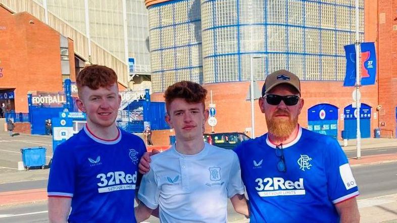 Three men looking at camera, two wearing blue Rangers FC tops, one with a ginger beard, cap and sunglasses, in front of Rangers' football stadium