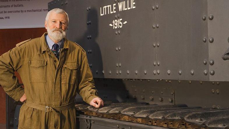 A man with white hair and a beard wearing pale brown overalls standing next to a large black tank.