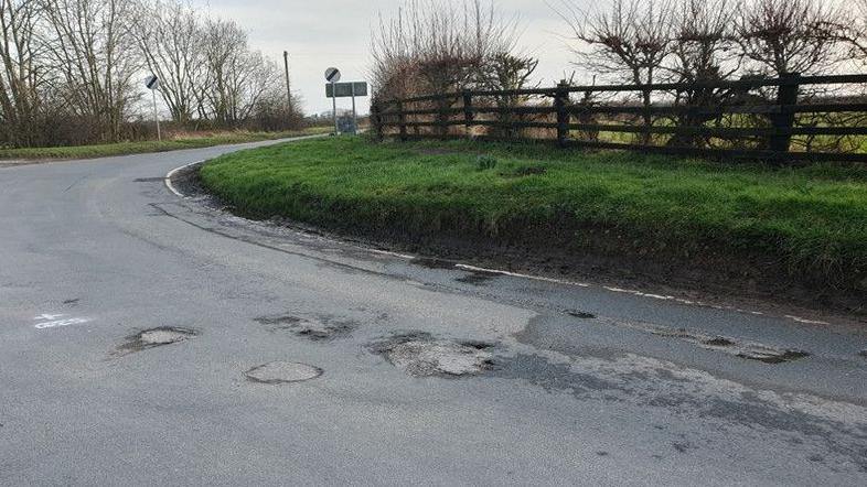 A well-worn section of rural road between Brandesburton and North Frodingham, showing evidence of potholes and patching work. Next to the road is a grass verge and hedgerow.