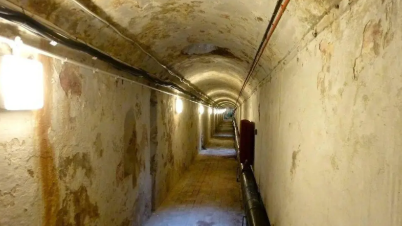 A tunnel beneath the Royal Pavilion in Brighton. The tunnel has straight side and a curved roof. The walls are white but the paint is peeling away. There is a square light on the left wall.