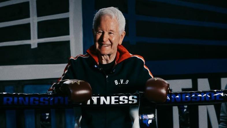 Frank Gilfeather standing inside a boxing ring wearing a dark tracksuit with a red and white trim, he is also wearing brown boxing gloves with golden crowns on them.