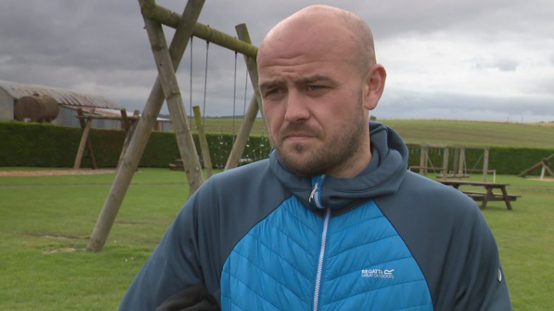 A man with a blue jacket and a beard stands outside in a playpark