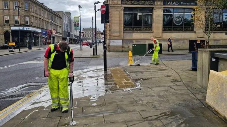 Two men in hi-vis clothing using power washers to clean streets of gum.