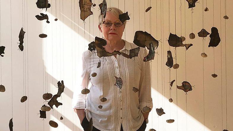 Sara Davidmann wearing a white shirt, glasses and black trousers standing behind burnt photo fragments and stones hanging from the ceiling on strings