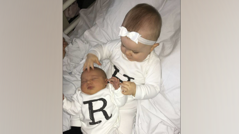 Harper, a young toddler, sits on a hospital bed holding newborn baby Romie. They are both wearing white baby grows with their initials on the front in black lettering. 