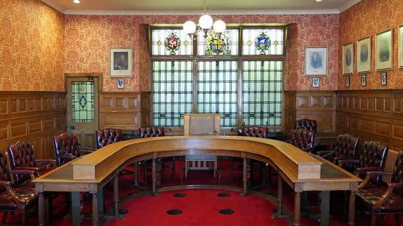 The Legislative Council Chamber, which has a large horseshoe shaped table in the middle surrounded by maroon coloured padded leather chairs. The room had wooden panelling on the walls and a red carpet, and there is a large window at the far end which has panels containing crests in stained glass at the top.