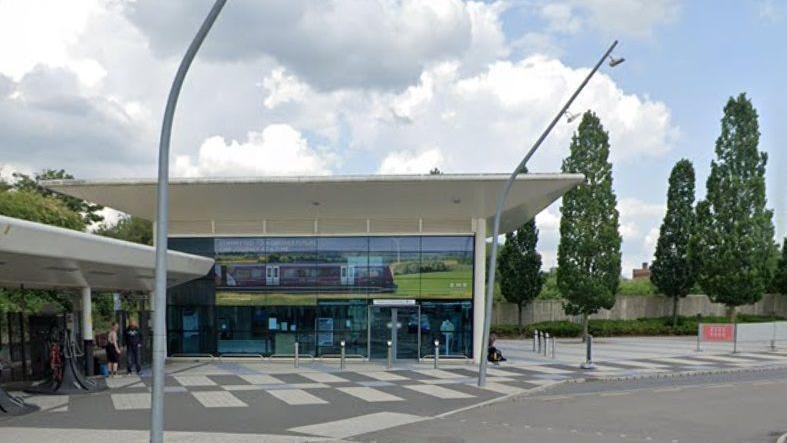 Corby station - a single-storey, glass-fronted building with a white roof. There is a white-roofed shelter to the left and trees to the right.