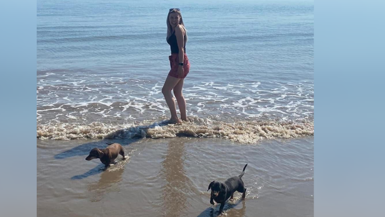 Josie Smith, who has long blonde hair and sunglasses on her head, is wearing red shorts and a black vest top and standing on the beach with her feet in the sea. The blue sea and sky are behind her, while in front of her on the sand are two dachshunds, one tan and one black. 