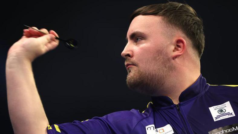 Luke Littler is aiming a dart in his right hand, he is wearing a purple top with sponsors on it, the background is black.