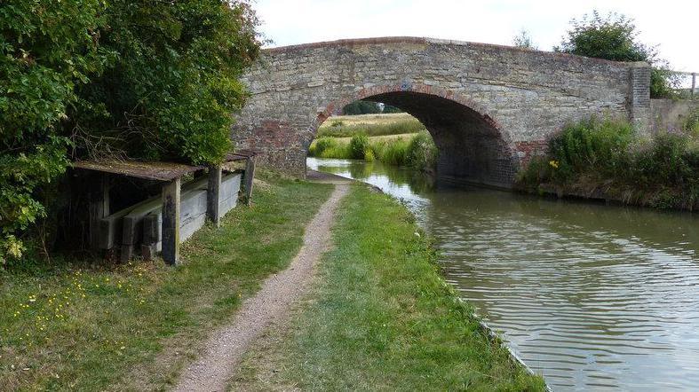 An old stone bridge arches over a canal, with a towpath running alongside it.