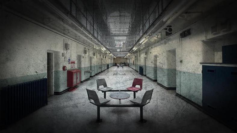 Four chairs and a small table are placed in the long hallway of Shepton Mallet prison. There is a net which separates the ground floor with the other floors.