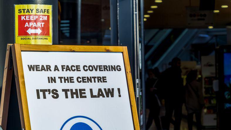 A business in a shopping centre displays a sign asking people to wear a face covering during the Covid pandemic. A further sign reminds people to "stay safe" and follow social distancing rules and keep two metres apart.