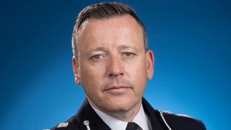 A close-up photo of the acting chief constable of West Midlands Police. He wears a black police uniform and stands in front of a blue background, looking into the camera.