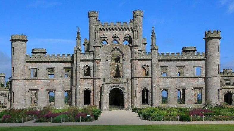 The entrance of Lowther Castle. The large stone building with towers is surrounded by grassland and gardens. 