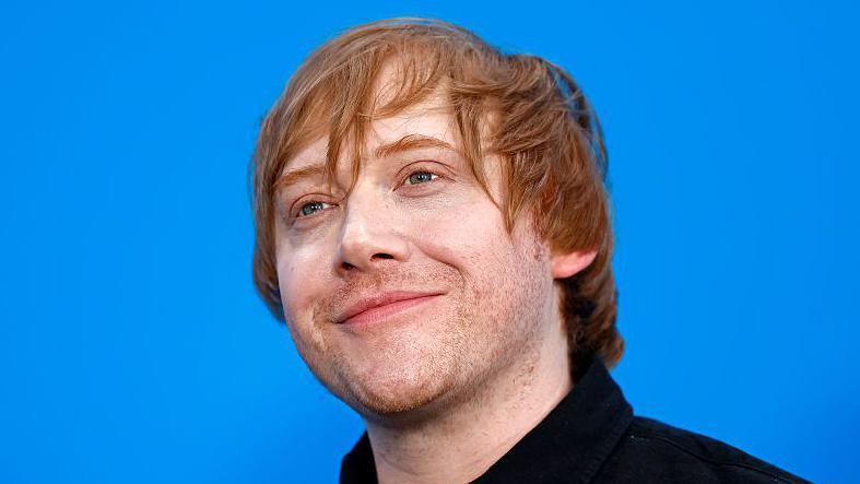 Rupert Grint is stood against a blue background and smiling. He has auburn hair and is wearing a black shirt. 