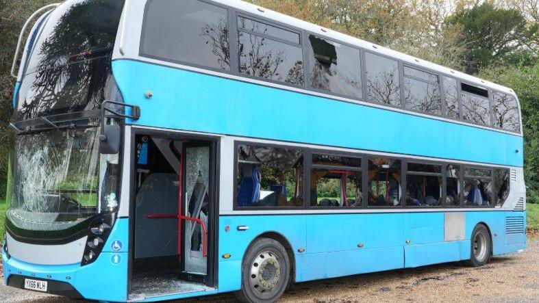 A light blue double-decker bus with smashed windows