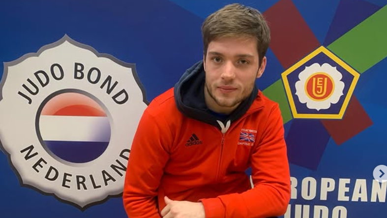 Rowan wearing a red GB zipped top with an Adidas sponsor logo. He is in front of a board that reads Judo Bond, Netherlands and has the European Judo logo on it which is red, green and blue around a white flower and yellow pentagon.