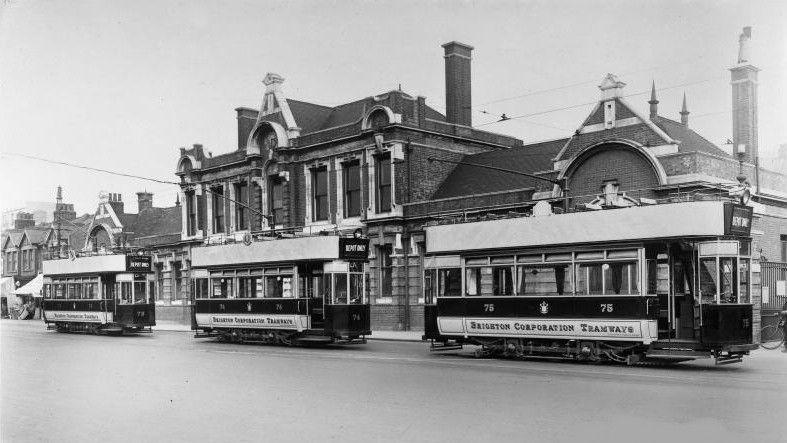 Black and white image of three trams parked on a roadside in front of a large brick building