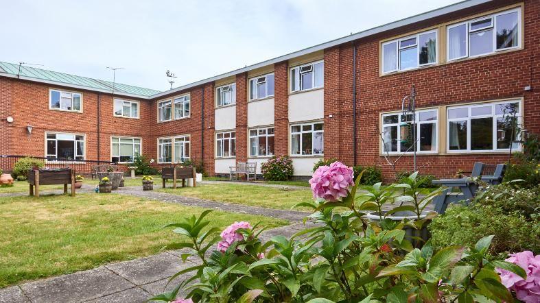 Linden Court in Watton; a red brick building with some cream-coloured panels around the central windows. It has a green copper sheet roof, with lawns and pink hydrangeas in the garden, along with some benches and seating.