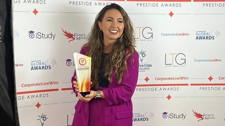 Lissy Cleminson. She is pictured holding an award. She has long dark hair and is wearing a purple suit with a black top. She is smiling, looking to the side of the camera. 