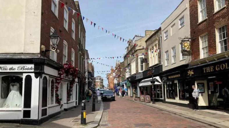 A wide shot of Rochester High Street on a sunny day with bunting across the street and shops.