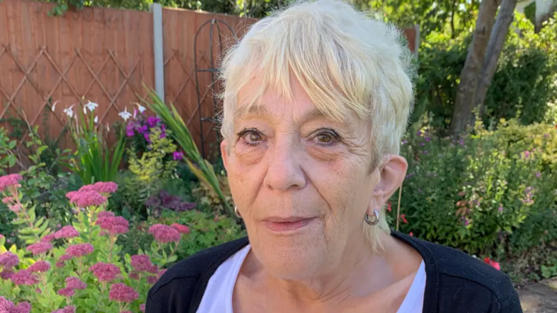 A picture of the head and shoulders of a woman with blond hair with the fence, trees and flowers of her garden behind her.