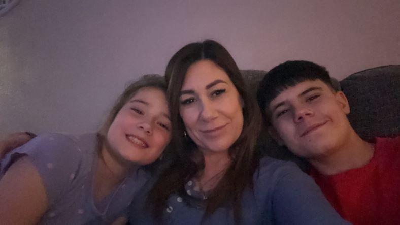 Sian Deeks sat on a sofa with long brown hair smiling with two children. A girl is sat on her left leaning in and smiling and a boy with black short hair and a red top is also leaning in and smiling.