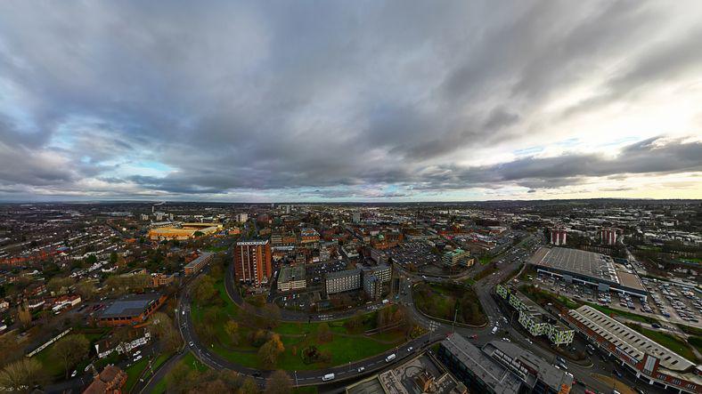 An aerial view of Wolverhampton. There are lots of buildings and the skyline goes for miles. It is a cloudy day. The yellow stadium of Wolverhampton Wanderers can be seen. 