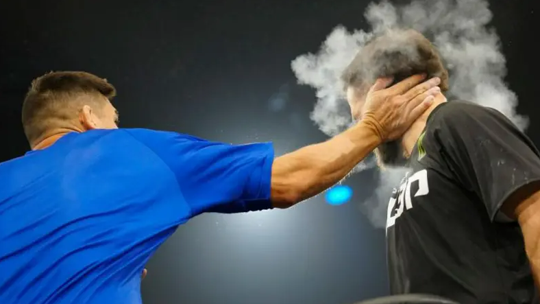 A man in a blue t-shirt slapping a man in a black t-shirt with white writing on it. He is slapping him with his right hand. A cloud of chalk dust and a man's hand is obscuring the other man's face.