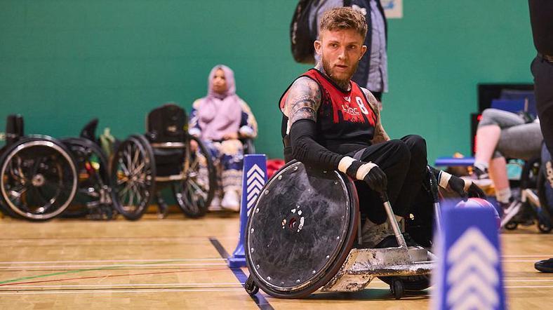 Joel Connor-Saunders covered in tattoos, wearing a red tank top and playing wheelchair rugby. There is a wood floor, green wall and people out of focus watching in the background.